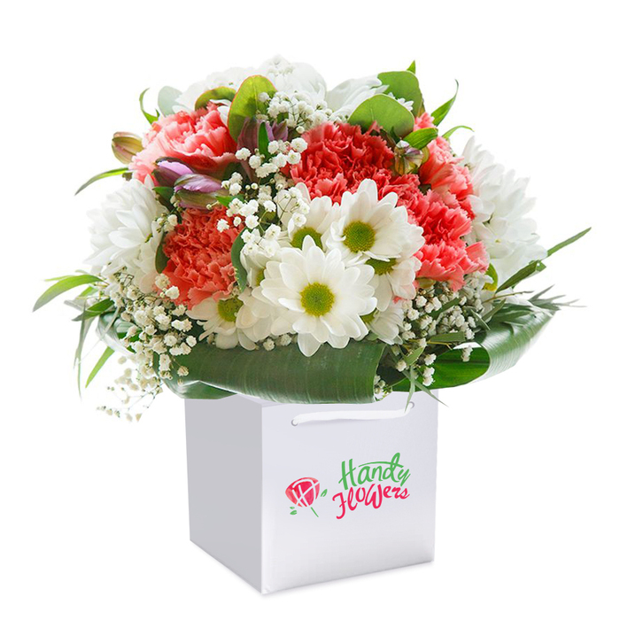 In this artfully composed bouquet, Pentonville's distinctive style comes alive through a rich orchestration of colour and form. Soft coral-pink and striking red carnations, each bloom full with abundant, ruffled petals, rise in a rounded arrangement from a modern white gift bag. Scattered between these exuberant blooms, crisp white daisies with vibrant lime-green centers offer cheer and contrast, evoking the brightness of a sunny morning at Joseph Grimaldi Park. A fine veil of delicate white gypsophila adds touches of airiness and light movement, harmonising the palette with its gentle texture. Hidden within, the subtler beauty of purple-tipped, unopened buds lends tantalising depth and a sense of stories still unfolding. Glossy, deep green leaves form a strong, graceful collar around the bouquet's base, their texture and sheen grounding the ensemble. Practical ribbon handles and minimalist bag design ensure that the arrangement is ready for delivery-whether it will grace a flat along Caledonian Road or be gifted at a gathering nearby. Set against a pure white background, the vibrant hues and florist's skill shine through, making this bouquet an ideal centrepiece for life's special milestones in Pentonville.