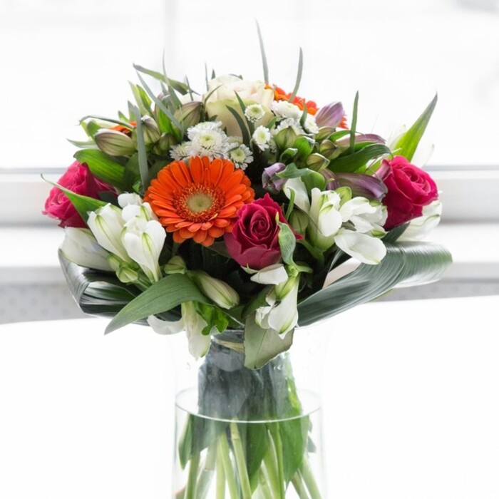 This elegantly composed bouquet, echoing the artistry found in Pentonville's vibrant floral scene, is a showcase of colour harmony and freshness. The focal point is a cluster of gerbera daisies-two vivid orange, two softly blushing peach-each with deep brown centre discs that add drama and sophistication. Flanking these, lushly furled magenta roses project romance and refinement, while bright white alstroemeria and petite white spray roses interrupt the palette with splashes of clarity and light. Hints of lighter pink blooms are nestled throughout, building a sense of fullness and natural abundance that recalls a garden at its peak, perhaps inspired by walks to the nearby Regent's Canal. Wide, curved aspidistra leaves, deep and glossy, are arranged in a sculpted collar around the bouquet's base, lending structure, contrast, and vibrancy reminiscent of Pentonville's leafy pockets. Fine, pointed greenery delicately fills the gaps, further enhancing the arrangement's round, generous form. The bouquet rests in a tall, crystal-clear glass vase, with stems arching elegantly downward-testament to the freshness Florist Pentonville is known for. Gently diffused daylight bathes the flowers and foliage, drawing out every nuance of tone and texture without distraction. Perfect for commemorating achievements, sending thanks, or gracing a contemporary home near Islington, this arrangement brims with warmth, artistry, and the personal touch of a Pentonville florist.