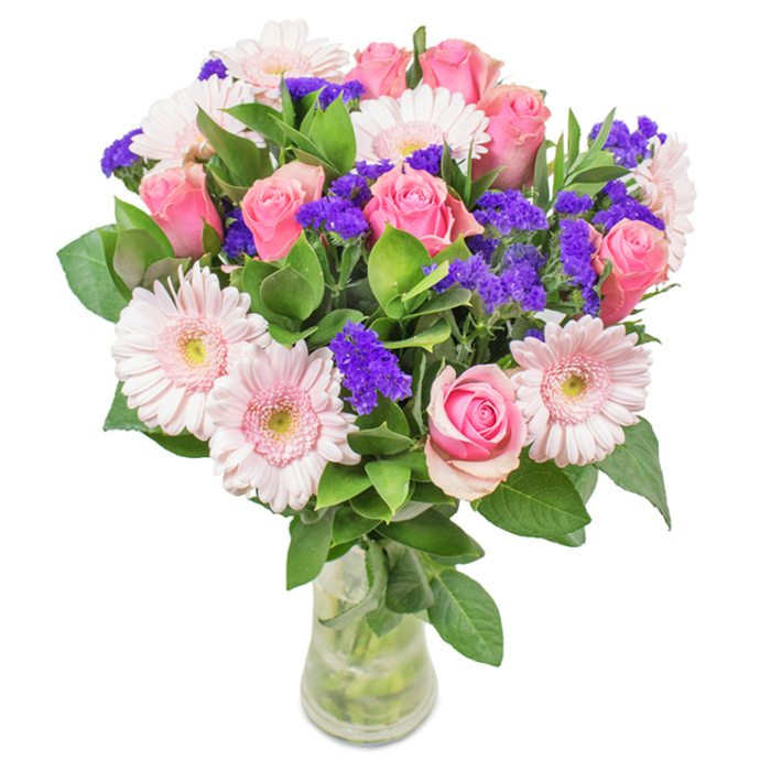 Visually striking against a crisp white backdrop, this floral arrangement is composed in a clear glass vase, the waterline and green stems visible beneath a domed canopy of blooms. Centrally featured are approximately seven to nine medium-pink roses, some still in bud while others gently unfurl in the classic spiral formation, each petal layered in soft gradients. Intertwined with these are five to seven pale gerberas-large, round flower heads with lighter, nearly white, central discs and a halo of blush pink petals. Between the main blooms, clusters of vibrant purple statice provide a textured, contrasting accent, infusing the bouquet with a lively touch reminiscent of the vibrant scene at Exmouth Market. Enveloping the arrangement, lush, emerald green foliage adds a sense of fullness and freshness. The overall composition, luminous and balanced, suggests a freshly delivered gift-equally suited for Pentonville offices, birthdays, sweet anniversaries, or simply to grace a space with cheerfulness and style. The bouquet's interplay of colour and form echoes local creativity, conveying joy, connection, and refined taste.
